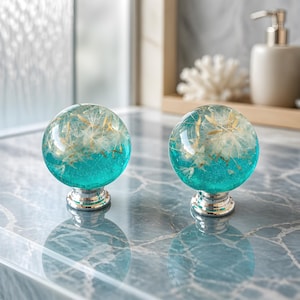 May include: Two decorative cabinet knobs. Each knob features a clear, spherical resin design with a turquoise base and embedded dandelion seeds. The knobs have a silver-colored base and are displayed on a marble surface.