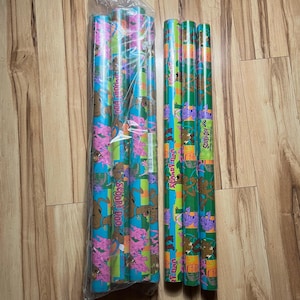 Könnte beinhalten: Mehrere Rollen buntes Geschenkpapier mit Scooby-Doo-Figuren. Das Papier hat ein lebendiges Design mit blauem, grünem und lila Hintergrund. Die Rollen sind gebündelt.