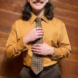 May include: A mustard yellow button-down shirt with a brown patterned tie. The tie features a repeating pattern of brown circles and squares. The shirt is paired with brown pants and a brown belt.