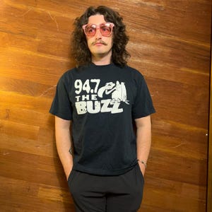 May include: A black short-sleeved t-shirt featuring a white graphic print that reads 94.7 THE BUZZ with an illustration of a bird. The shirt is paired with black trousers and pink-tinted sunglasses, creating a monochromatic look with a pop of colour.