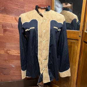 May include: A long-sleeved shirt with a tan suede-like yoke and trim, and a navy blue body. The shirt has a mandarin collar, button closures, and decorative accents on the chest pockets. The shirt is displayed on a mannequin.