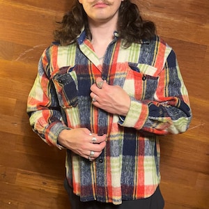 May include: A colorful plaid button-up shirt in a mix of red, navy, green, and cream. The shirt has two chest pockets and a classic collar. The fabric appears to be a thick, warm material, suitable for cooler weather.