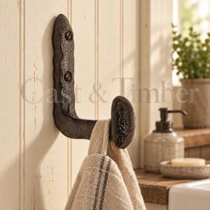 May include: A dark, textured, L-shaped metal wall hook with a rough, cast-iron appearance. The hook is mounted to a vertical surface with two visible screws. A beige, woven fabric towel with a thin dark stripe hangs from the hook's curved end.
