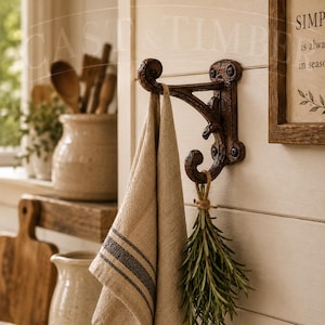 May include: A dark brown, ornate cast iron wall hook with a double-pronged design mounted on a white wooden wall. A beige linen towel with a dark stripe hangs from the top hook, and a bundle of fresh green rosemary is tied to the bottom hook.