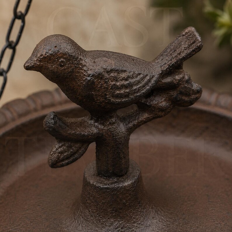 Op de afbeelding: Een donkerbruin gietijzeren vogelbeeldje zittend op een tak, bevestigd in het midden van een ronde, gegroefde schaal. De vogel heeft een getextureerd oppervlak en staat boven een voetstuk, met een metalen ketting zichtbaar in de linkerbovenhoek.