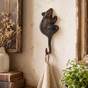 May include: A dark brown, cast iron wall hook shaped like a climbing mouse. The mouse figure is mounted to the wall with two visible screws, and its long, curved tail serves as the hook, currently holding a beige, textured fabric towel.