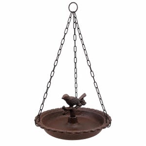 Op de afbeelding: Een hangende vogelvoederbak van donkerbruin gietijzer met een ronde schaal met een geschulpte rand en een centrale paal met een kleine vogel op een tak. De voederbak wordt opgehangen aan drie dunne zwarte metalen kettingen aan een ring.