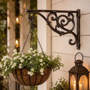 May include: A black metal wall bracket with ornate scrollwork supports a hanging basket filled with small white flowers. The basket is suspended by three thin black chains attached to a central hook on the bracket, featuring a natural fiber liner.