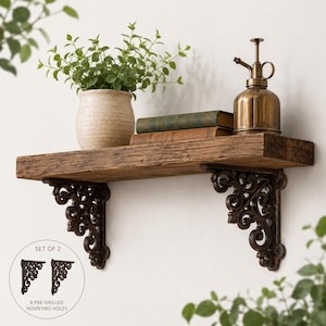 May include: A rustic wooden wall shelf supported by two dark brown metal brackets with ornate scrollwork. The shelf holds a potted green plant, two stacked books, and a brass-colored metal mister. A circular inset shows a set of two brackets.