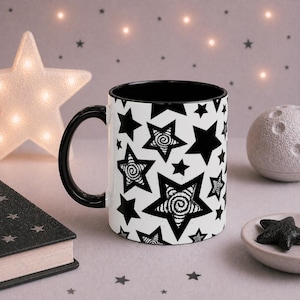 Puede incluir: Una taza de cerámica blanca con interior y asa negros, decorada con un patrón de estrellas negras. Algunas estrellas tienen un diseño en espiral. La taza está sobre una superficie clara con objetos decorativos.