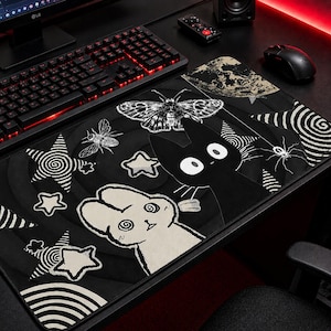 May include: Black mouse pad with a whimsical design featuring a black cat, a white rabbit, stars, a moth, and an insect. The pad has a black background with white and gray accents, and is placed on a desk with a keyboard and mouse.