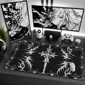 Pode incluir: Um tapete de secretária retangular preto com um design de cruz gótica branca intrincada, rodeado por padrões espinhosos semelhantes a videiras e motivos florais. O tapete está sobre uma secretária em frente a dois monitores com arte anime.