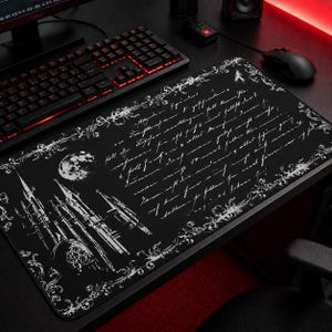 May include: Black rectangular gaming mousepad with a white gothic castle illustration, moon, and script. The mousepad has a decorative white border and is placed on a black desk with a keyboard and mouse.