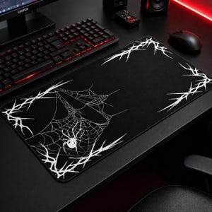 May include: Black rectangular mouse pad featuring a white spider web design with a spider in the center. The pad has a white spiky border. The pad is on a black desk with a keyboard, mouse, and other computer accessories.