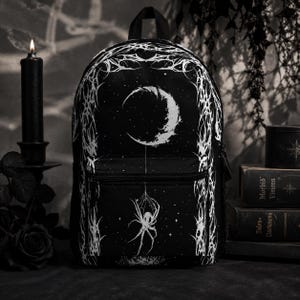 May include: A black backpack featuring a white graphic print of a crescent moon, a spider hanging from a web, and intricate thorny vine patterns along the edges. The bag includes a front zippered pocket and a main compartment with a top handle.