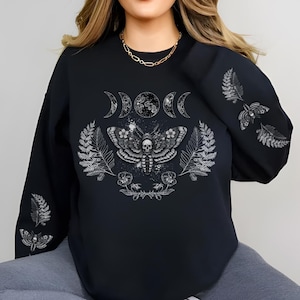 Puede incluir: Sudadera negra con un diseño detallado de polilla con una calavera, rodeada de helechos, flores y fases lunares. Las mangas también tienen ilustraciones de polillas y helechos. El diseño es en tonos grises y blancos.