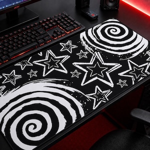 Op de afbeelding: Zwart-wit gaming muismat met een sterren- en spiraalontwerp. De mat heeft een zwarte achtergrond met witte sterren en spiraalpatronen. Een toetsenbord en computermonitor zijn zichtbaar.