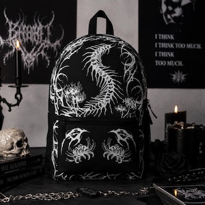 May include: A black backpack featuring a white graphic print of a spinal column and spider lily flowers. The bag has a top handle, a main zippered compartment, and a smaller front zippered pocket, all decorated with intricate, sharp-lined patterns.