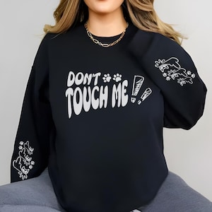 Puede incluir: Sudadera negra con el texto blanco "DON'T TOUCH ME!". La sudadera presenta huellas de patas e ilustraciones de gatos en las mangas. Se ve un collar de cadena dorada.