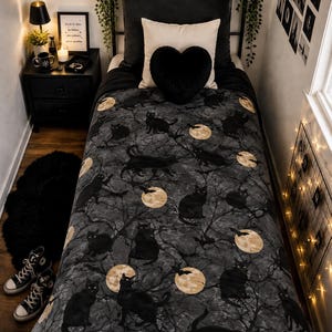 May include: A twin-sized bed featuring a dark gray comforter with a repeating pattern of black cats, bare tree branches, and full moons. The bed includes a black headboard, a cream-colored square pillow, and a black heart-shaped plush pillow.