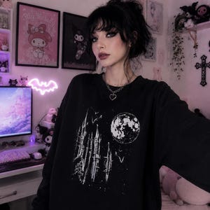 May include: Black sweatshirt featuring a white graphic of a gothic cathedral, a full moon, and flying bats. The design is a distressed, vintage style. The sweatshirt is a crew neck style.