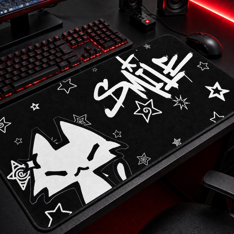 May include: Black rectangular mousepad with white graphic art. The design includes a stylized cat face, stars, and the word "Smile" in a graffiti-style font. The mousepad is on a black desk with a keyboard and mouse.