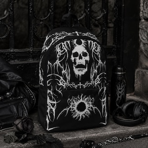 May include: A black backpack featuring a white graphic print of a skull and a circular sun-like emblem surrounded by intricate, sharp, thorn-like patterns. The design includes small cross symbols above the skull and within the decorative motifs.