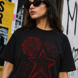 以下が含まれることがあります： 黒の半袖Tシャツで、かすれた赤色のグラフィックプリントが施されています。デザインは、大きな渦巻き模様、渦巻きの中心を持つ目立つ五芒星、そしてその周囲に散らばるいくつかの小さな星で構成されています。