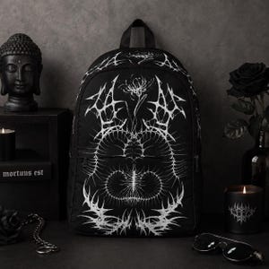 Puede incluir: Una mochila negra con un estampado gráfico blanco, complejo y simétrico, de patrones espinosos tipo telaraña y un diseño floral central. La mochila cuenta con un asa superior, un compartimento principal con cremallera y un bolsillo frontal.