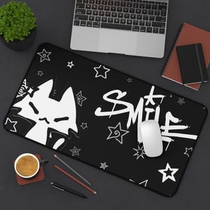 May include: Black rectangular desk mat featuring a white cat graphic and the word "Smile" in a graffiti-style font. White stars are scattered across the mat. A laptop, mouse, and other office supplies are visible.
