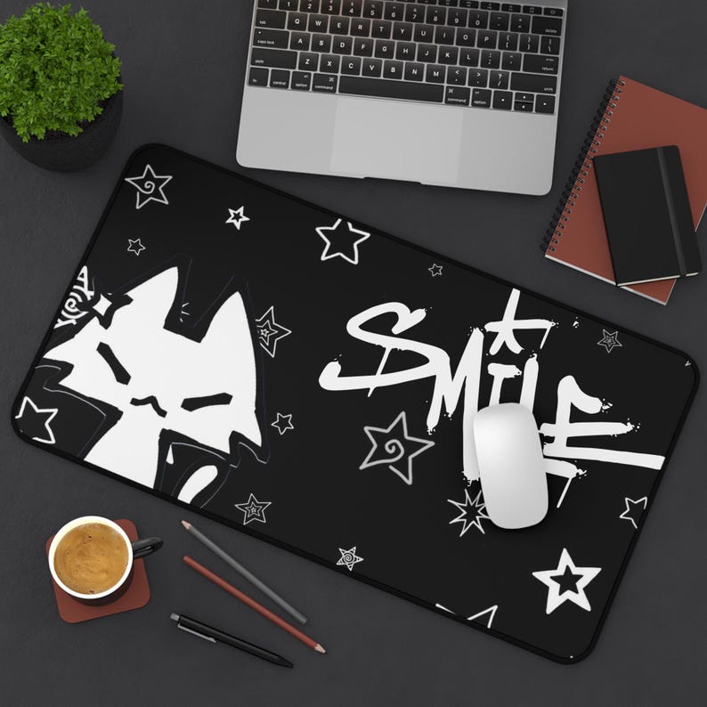 May include: Black rectangular desk mat featuring a white cat graphic and the word "Smile" in a graffiti-style font. White stars are scattered across the mat. A laptop, mouse, and other office supplies are visible.
