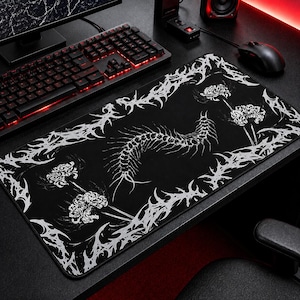 Puede incluir: Alfombrilla de ratón rectangular negra con un diseño blanco estilizado. El diseño presenta un elemento central en forma de espina dorsal, detalles florales y un borde decorativo. La alfombrilla está sobre un escritorio negro con teclado y ratón.