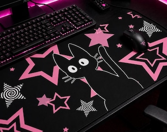 May include: Black desk mat featuring a cartoon cat illustration and pink star patterns. The mat has a black cat with white eyes and whiskers, surrounded by pink stars and spiral designs. A keyboard and mouse are on the desk.