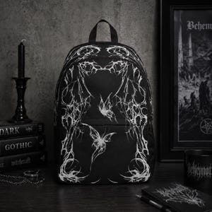 Puede incluir: Una mochila negra con un intrincado patrón gráfico blanco similar a una telaraña, con dos ilustraciones de mariposas centradas en la parte frontal. El diseño incluye líneas finas y afiladas que se extienden por la superficie de la tela.