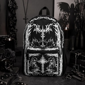 May include: A black backpack featuring a white, intricate, symmetrical tribal-style graphic print. The design includes sharp, thorny, and vine-like patterns covering the main compartment and the front zippered pocket, which displays a central cross.