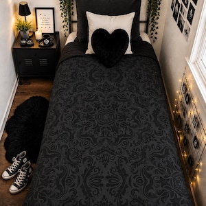 May include: A twin bed featuring a black duvet cover with a dark gray damask pattern. The bedding includes a large black rectangular pillow, a white square pillow, and a plush black heart-shaped cushion centered on the bed surface.