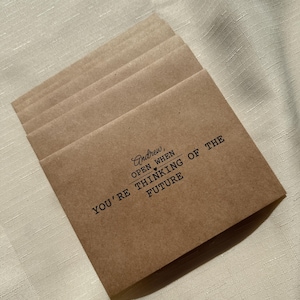 May include: A stack of brown paper envelopes with the text "Andrew, OPEN WHEN YOU'RE THINKING OF THE FUTURE" printed in black. The envelopes are arranged on a cream-colored fabric surface.