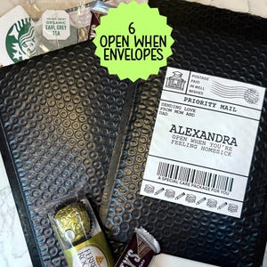 May include: Two black bubble mailers with a lime green starburst label reading 6 OPEN WHEN ENVELOPES. One mailer has a white shipping label addressed to ALEXANDRA, OPEN WHEN YOU'RE FEELING HOMESICK, with a Starbucks card, tea, and chocolates nearby.