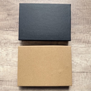 May include: Two rectangular boxes are positioned one above the other. The top box is solid black with a smooth, matte finish. The bottom box is a natural light brown kraft paper color with a similar smooth, matte texture and clean edges.