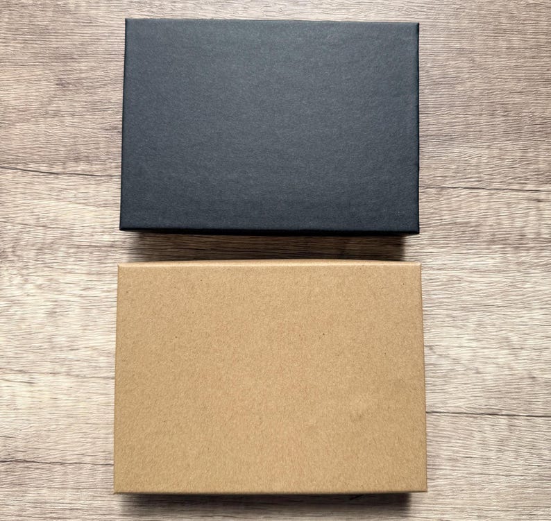 May include: Two rectangular boxes are positioned one above the other. The top box is solid black with a smooth, matte finish. The bottom box is a natural light brown kraft paper color with a similar smooth, matte texture and clean edges.