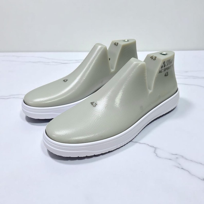 Op de afbeelding: Twee grijze schoenmallen met witte zolen. Elke mal is gemarkeerd met het nummer "43" en de tekst "AC SPORT". Deze mallen worden gebruikt om schoenen te vormen en worden getoond op een wit marmeren oppervlak.