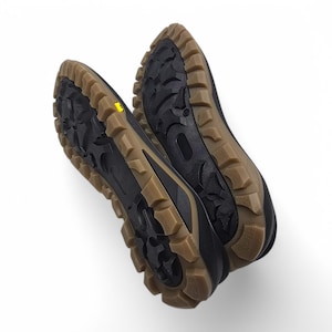 May include: A pair of black and brown hiking shoes, soles facing upwards. The shoes feature a rugged tread pattern with brown outsoles and black uppers. A small yellow accent is visible on one sole. Designed for outdoor activities.