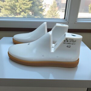 May include: Two white shoe lasts with brown soles are displayed on a white surface. The lasts have a curved shape, with the text "ITAL STOP AC LIKE 42 Y" and the number "42" printed on the side. The lasts are for shoe making.