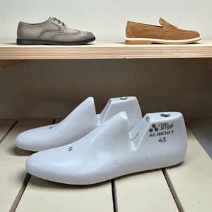 Puede incluir: Un par de hormas de plástico blanco con el texto Ital Step AC RICHI T 43 impreso en el lateral, situadas bajo un zapato oxford de ante gris y un mocasín de ante tostado con suela blanca sobre un estante de madera.
