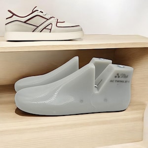 Könnte beinhalten: Hellgraue Schuhspanner mit dem Text "ITAL STEP AC TWINS 37 K" an der Seite, auf einem Holzregal. Darüber ein weißer Sneaker mit bordeauxfarbenen Akzenten.