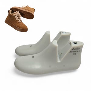 May include: A pair of light gray shoe forms with the number "39" printed on them. A brown suede boot with cream-colored laces is in the background. The shoe forms have a metal bar and the text "AC YUGA Y 39".