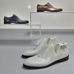 May include: Three pairs of men's dress shoes in blue, brown, and black, displayed on white shelves. Two white shoe lasts with black soles are in the foreground. The shoe lasts have the number 42 printed on them.