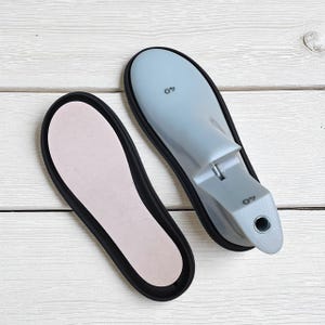 May include: Two shoe lasts and an insole are displayed on a white wooden surface. The shoe lasts are light blue and marked with the number "40". The insole is pink with a black border. The items are for shoemaking.