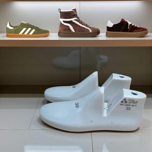 May include: A display of footwear, including green sneakers with white stripes, brown high-top boots with white trim, and burgundy sneakers. Two white shoe lasts with the number 39 are in the foreground.