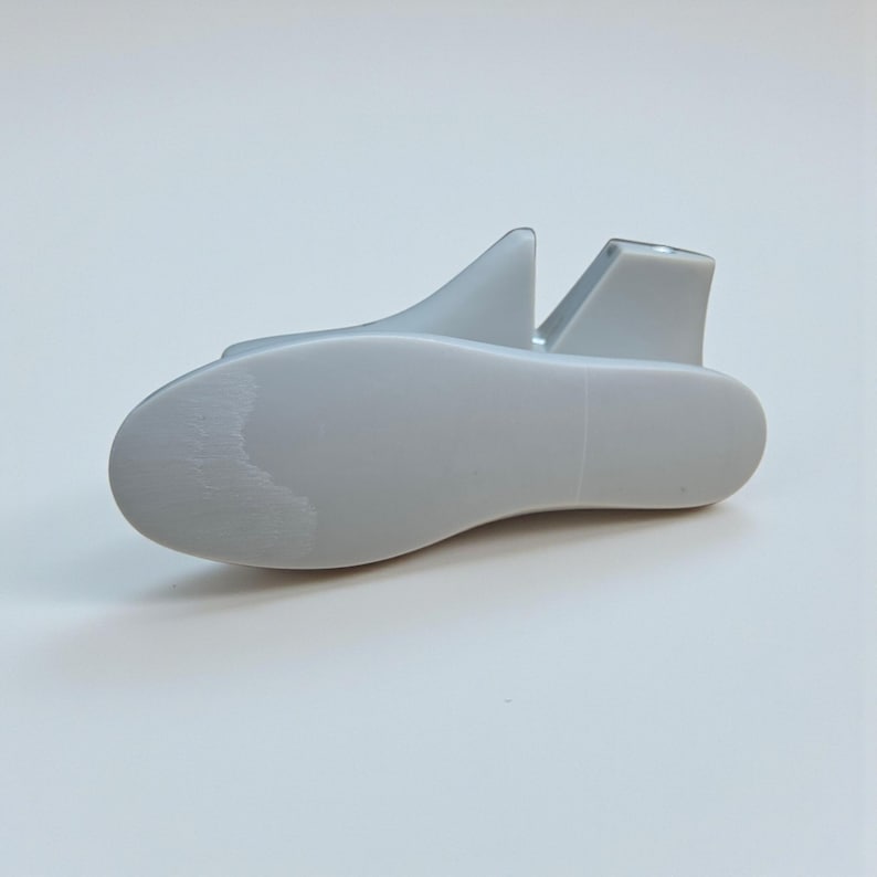 May include: A light gray shoe stretcher made of plastic. The shoe stretcher has a rounded toe and a heel support with two fins. The shoe stretcher is designed to maintain the shape of shoes and prevent creases.